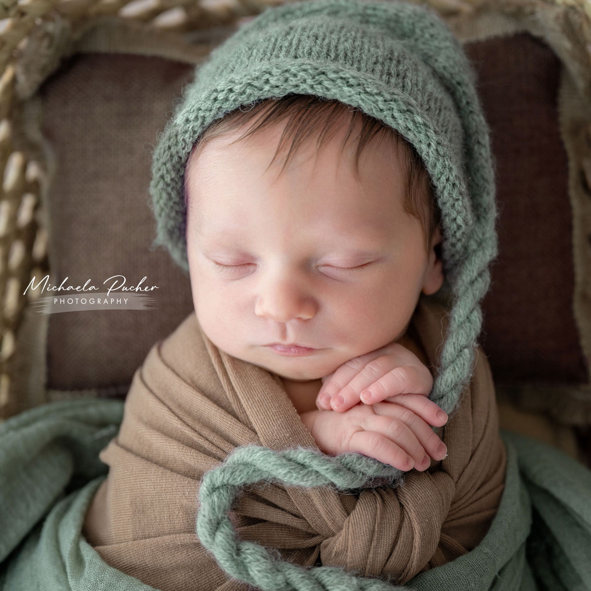 Schlafendes Neugeborenes, eingewickelt in braune und grüne Tücher, mit einer weichen grünen Strickhaube und gefalteten Händen.